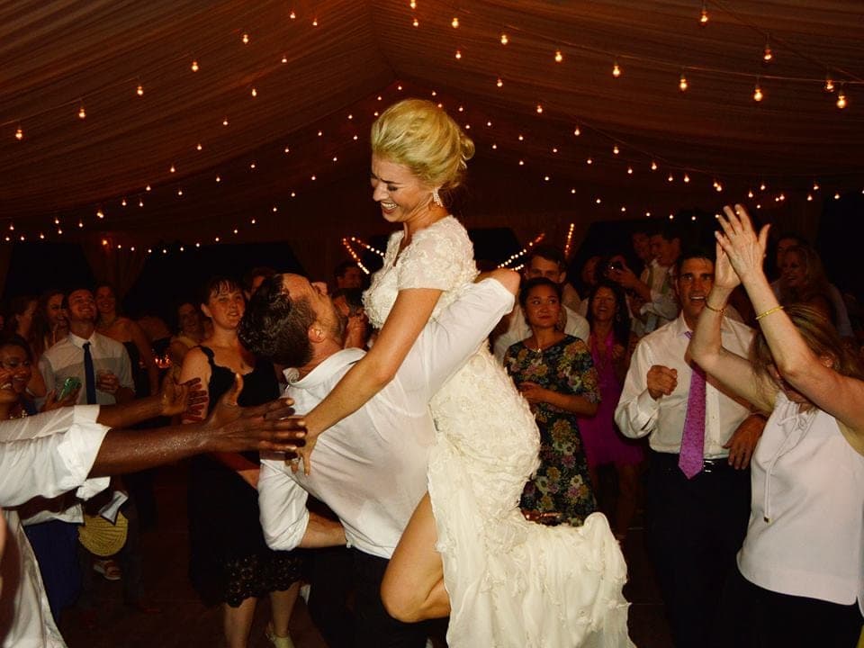 A bride lifted above a crowded dance floor under string lights
