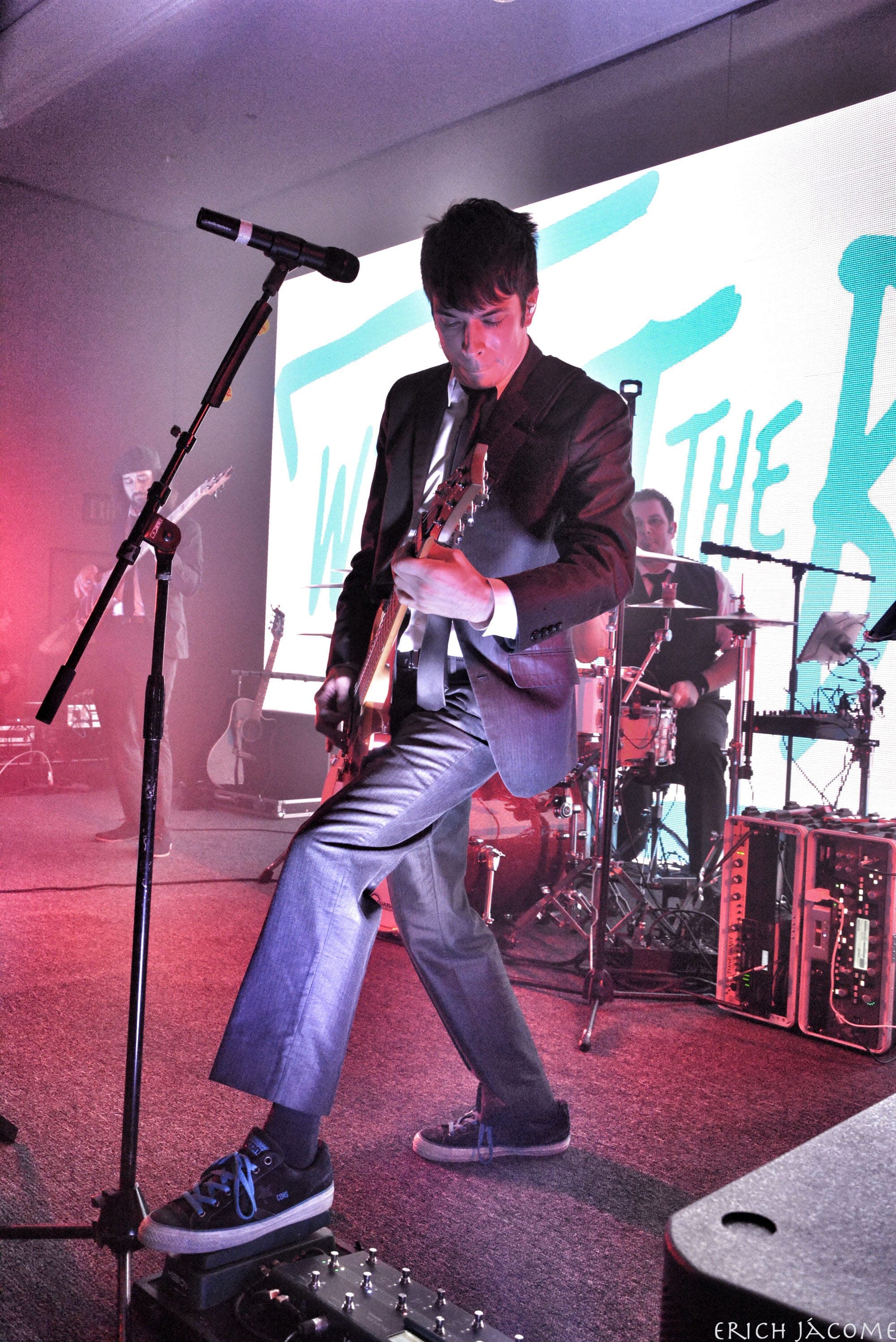 The band's guitarist mid-song in a grey suit, with the drum kit behind him