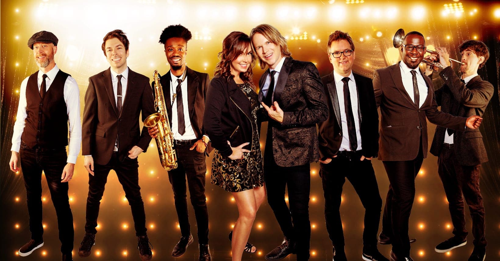 The band in black suits and dresses under warm string lights, holding a saxophone, a trumpet and guitars